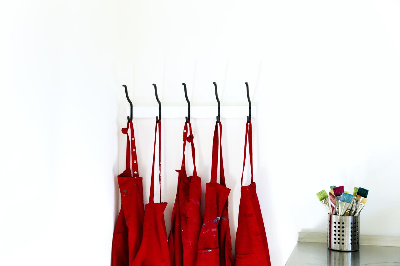 An fünf Garderobenhaken hängen vor einer weißen Wand rote Arbeitskittel. Rechts daneben steht ein Metalltopf mit bunten breiten Pinseln.