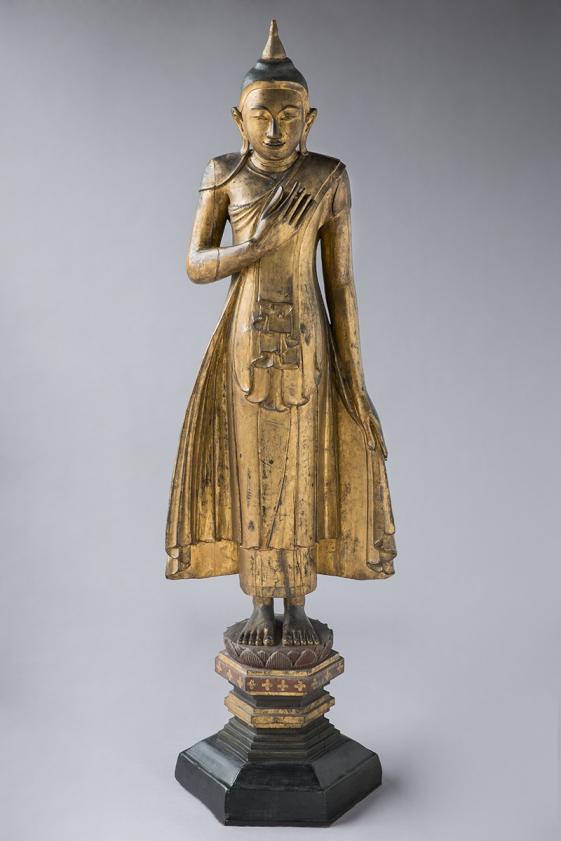 Buddha, Foto: Paul Schwimweg - © MARKK Farbfotografie einer stehenden goldenen Buddha Statue auf einem schwarzen Sockel. Die Figur hat die rechte Hand auf der Brustmitte platziert.
