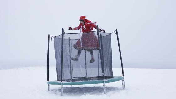Trambo 1 - ©Maja Helander Das Bild zeigt ein großes Trampolin in mitten einer Schneelandschaft. Eine Frau in roter Trachtenkleidung springt auf dem Trampolin, während es schneit.