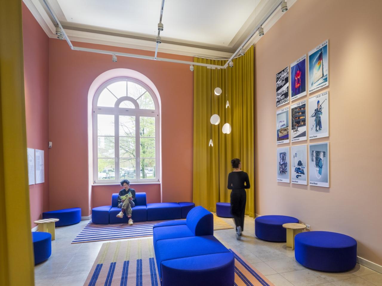 Zwei Besucher*innen in einem hohen, pfirsichfarben gestrichenen Raum mit azurblauen Sofas und großen Fenstern.