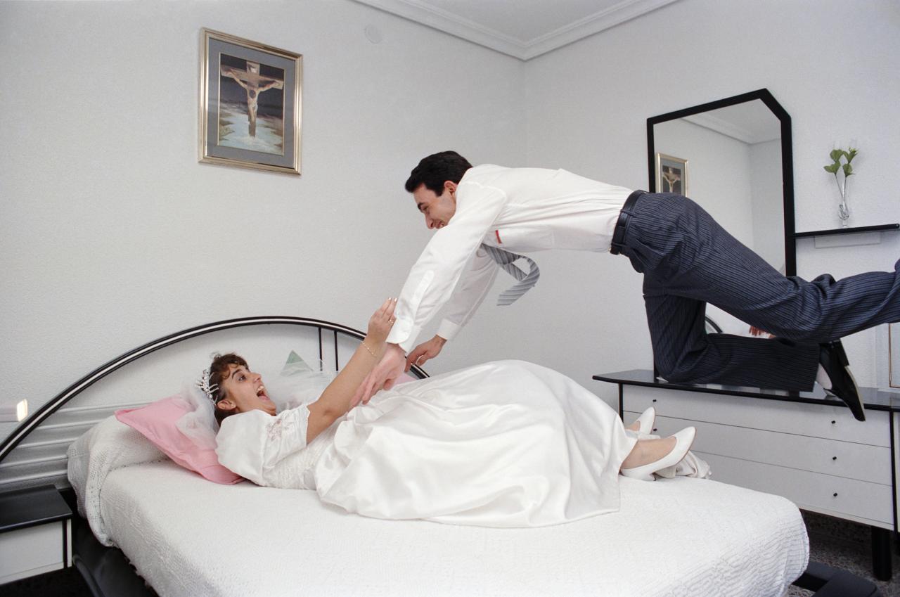 Zu sehen ist ein Ehepaar in einem Schlafzimmer. Die Braut liegt auf dem Bett und der Mann springt zu ihr. Beide freuen sich.