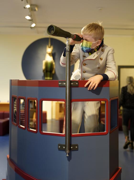 Kinder entdecken die Wunderkammer im Altonaer Museum. - © Kristine Thiemann Farbfotografie eines Jungen, der in Kapitänsverkleidung und mit buntem Mund Nasen Schutz auf einem nachgebauten Schiff durch ein Fernglas schaut.