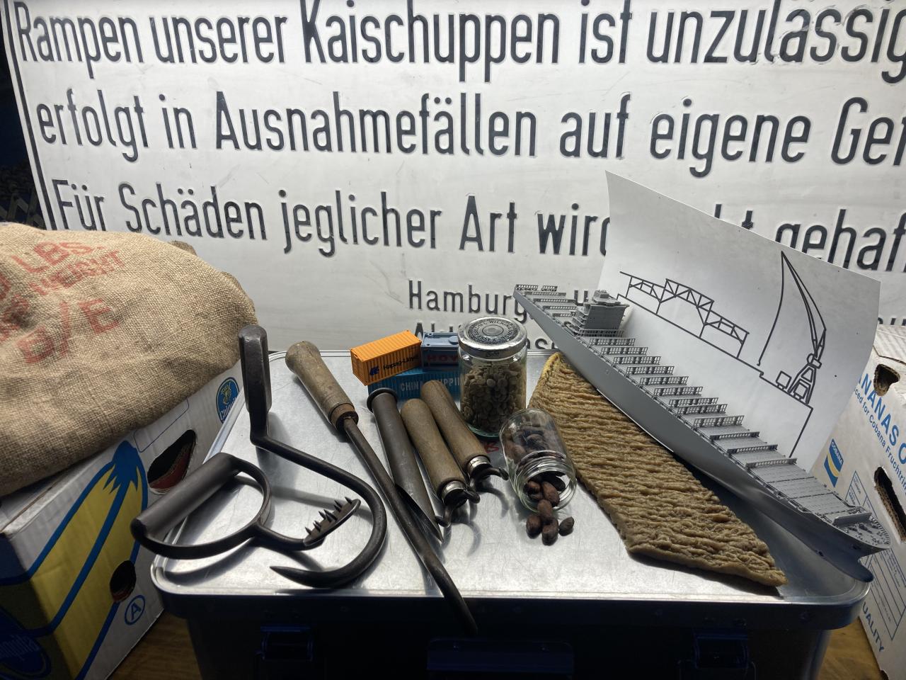 Das Foto zeigt einen Materialkoffer voller Objekte, die bei der Führung zum Einsatz kommen: Seesack, Griepen, Bananenkisten, Containermodelle, Geruchsproben von Kakao und Kaffeebohnen, ein graues Modellschiff und Schwellkopien.