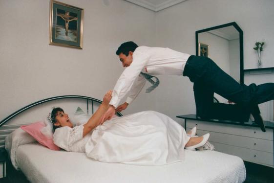 Zu sehen ist ein Ehepaar in einem Schlafzimmer. Die Braut liegt auf dem Bett und der Mann springt zu ihr. Beide freuen sich.  Juan de la Cruz Megias Mondéjar